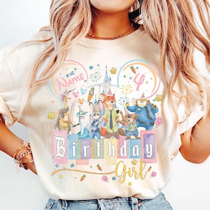 以下が含まれることがあります： クリーム色のTシャツ。カラフルな漫画のキャラクター、お城、そして「Birthday Girl」の文字が描かれています。デザインには数字の「4」と名前を入れるスペースがあり、星と花のアクセントが施されています。