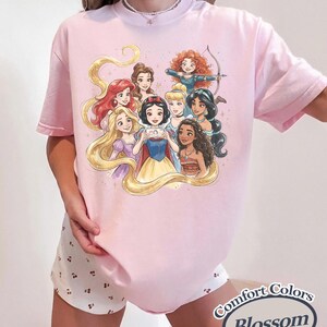 Op de afbeelding: Lichtroze T-shirt met een illustratie van tekenfilmprinsessen. De prinsessen worden in een groep afgebeeld, sommigen houden een telefoon vast. De woorden "Comfort Colors Blossom" staan rechtsonder op het shirt.