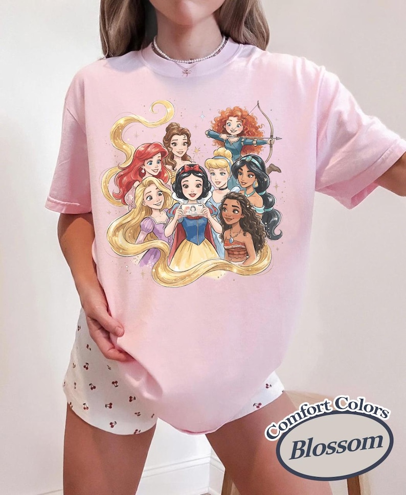 Op de afbeelding: Lichtroze T-shirt met een illustratie van tekenfilmprinsessen. De prinsessen worden in een groep afgebeeld, sommigen houden een telefoon vast. De woorden "Comfort Colors Blossom" staan rechtsonder op het shirt.