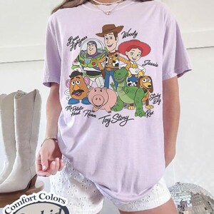 Puede incluir: Una camiseta lila Comfort Colors con un gr&aacute;fico de personajes de Toy Story, incluyendo a Woody, Buzz Lightyear y Jessie. La camiseta tiene el texto "Toy Story" debajo de los personajes y "Comfort Colors Orchid" en una etiqueta.