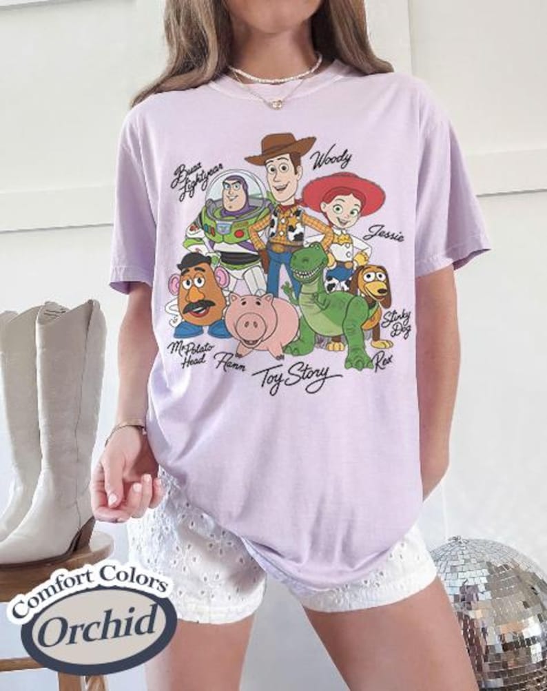 Puede incluir: Una camiseta lila Comfort Colors con un gr&aacute;fico de personajes de Toy Story, incluyendo a Woody, Buzz Lightyear y Jessie. La camiseta tiene el texto "Toy Story" debajo de los personajes y "Comfort Colors Orchid" en una etiqueta.