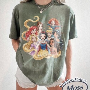 Op de afbeelding: Mosgroen Comfort Colors T-shirt met een afbeelding van geanimeerde prinsessen. Het ontwerp toont een groep prinsessen met lang haar en kleurrijke jurken. Het T-shirt heeft een relaxte pasvorm en korte mouwen.