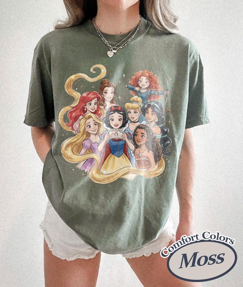 Op de afbeelding: Mosgroen Comfort Colors T-shirt met een afbeelding van geanimeerde prinsessen. Het ontwerp toont een groep prinsessen met lang haar en kleurrijke jurken. Het T-shirt heeft een relaxte pasvorm en korte mouwen.
