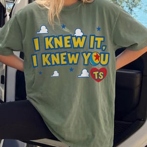 May include: An olive green short-sleeve t-shirt featuring the yellow text I KNEW IT, I KNEW YOU with a blue and yellow ball icon replacing the letter O. The design includes white cloud graphics, small blue stars, and a red heart containing the letters TS.