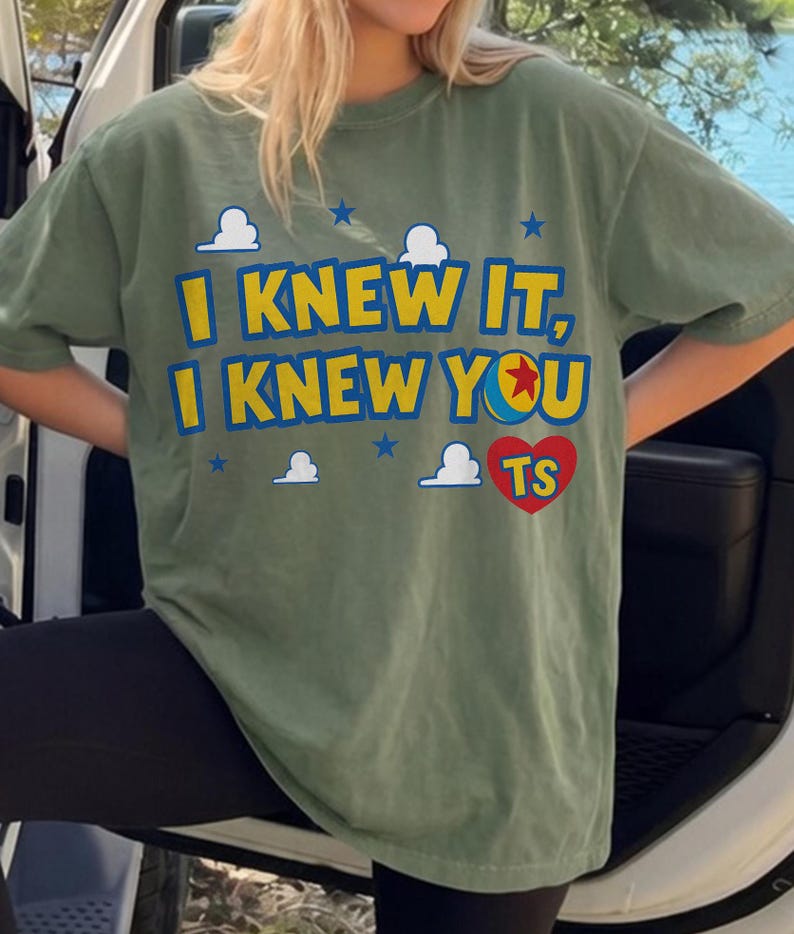 May include: An olive green short-sleeve t-shirt featuring the yellow text I KNEW IT, I KNEW YOU with a blue and yellow ball icon replacing the letter O. The design includes white cloud graphics, small blue stars, and a red heart containing the letters TS.