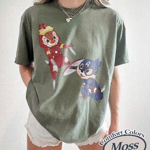 May include: Moss green t-shirt featuring cartoon characters dressed as superheroes. One character is in a red and gold Iron Man suit, and the other is in a Captain America costume. The shirt has a Comfort Colors label.