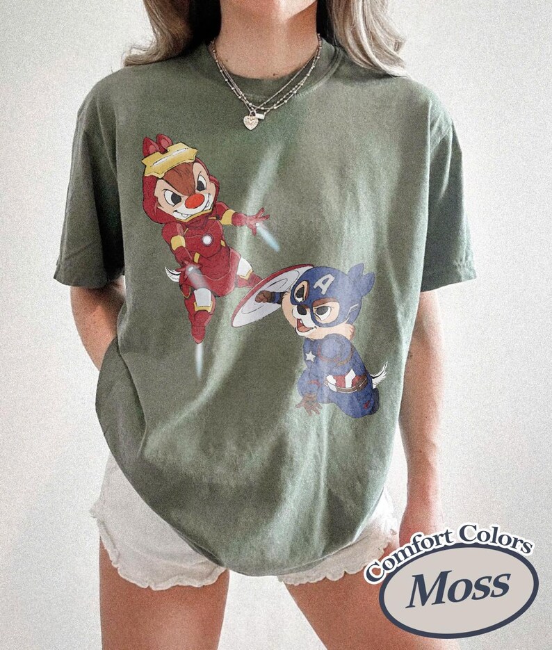 May include: Moss green t-shirt featuring cartoon characters dressed as superheroes. One character is in a red and gold Iron Man suit, and the other is in a Captain America costume. The shirt has a Comfort Colors label.