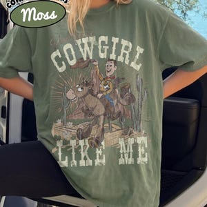 May include: A moss green short-sleeve t-shirt featuring a graphic print of a cartoon cowboy riding a horse in a desert setting with cacti. The text "COWGIRL LIKE ME" is printed in a distressed white font above and below the central illustration.