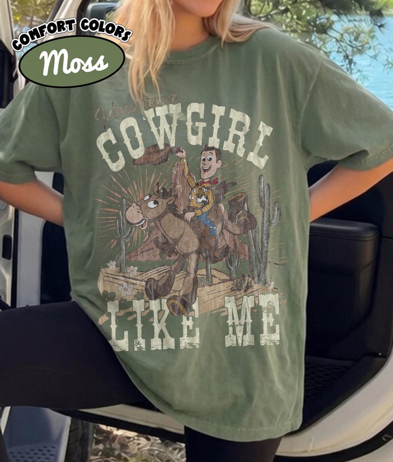 May include: A moss green short-sleeve t-shirt featuring a graphic print of a cartoon cowboy riding a horse in a desert setting with cacti. The text "COWGIRL LIKE ME" is printed in a distressed white font above and below the central illustration.