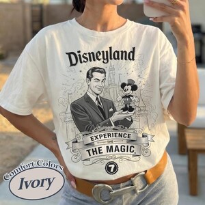 Pu&ograve; includere: T-shirt color avorio con grafica Disneyland. La grafica presenta un uomo che ammicca e tiene in mano una figura di Topolino, con la scritta "Disneyland Experience the Magic". La maglietta ha un'etichetta Comfort Colors.