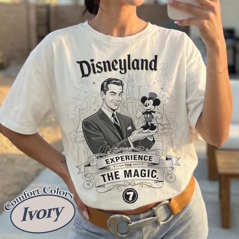 Pu&ograve; includere: T-shirt color avorio con grafica Disneyland. La grafica presenta un uomo che ammicca e tiene in mano una figura di Topolino, con la scritta "Disneyland Experience the Magic". La maglietta ha un'etichetta Comfort Colors.