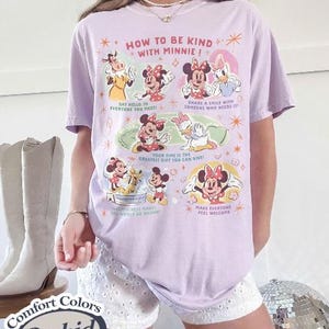 Puede incluir: Una camiseta lila Comfort Colors con un gr&aacute;fico de Minnie Mouse y sus amigos. La camiseta tiene el texto "HOW TO BE KIND WITH MINNIE!" y otras frases. La camiseta se combina con pantalones cortos blancos.