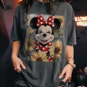 Puede incluir: Una camiseta de manga corta gris carb&oacute;n con un estampado gr&aacute;fico de Minnie Mouse con un vestido de lunares rojos y blancos y un lazo a juego, rodeada de girasoles amarillos. El dise&ntilde;o tiene una est&eacute;tica suave, deste&ntilde;ida y vintage.