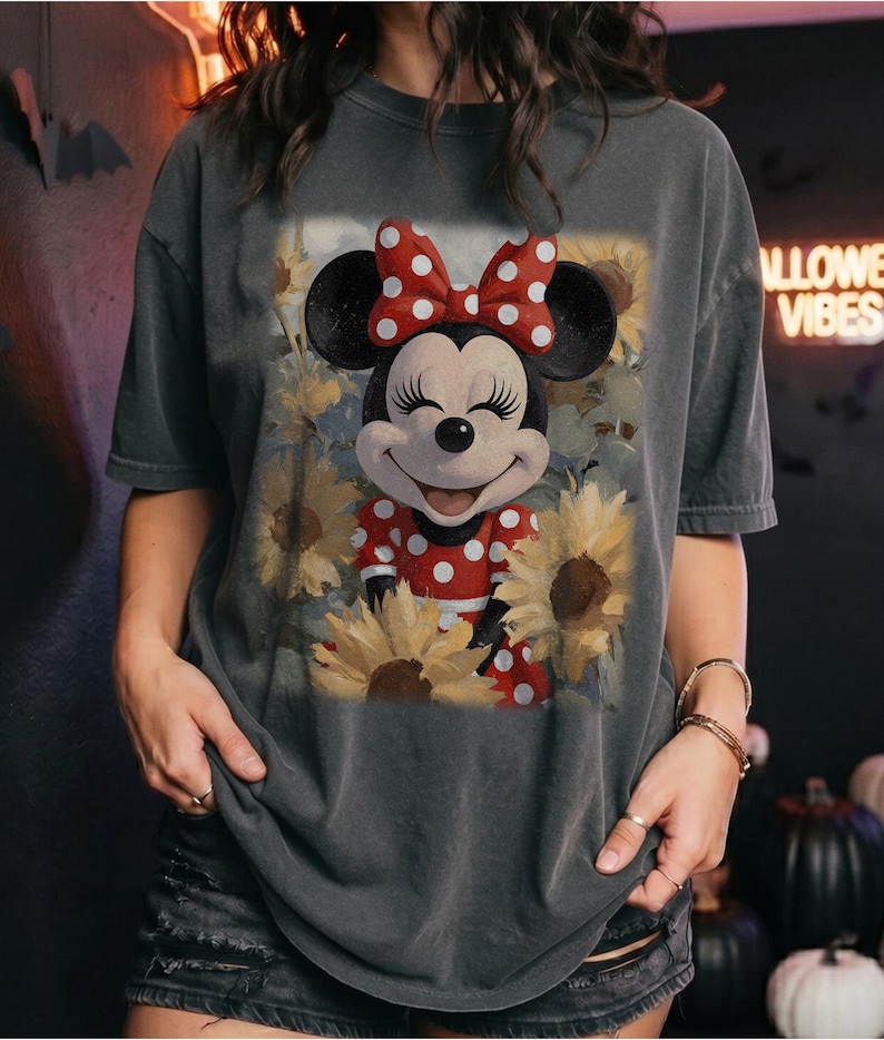 Puede incluir: Una camiseta de manga corta gris carb&oacute;n con un estampado gr&aacute;fico de Minnie Mouse con un vestido de lunares rojos y blancos y un lazo a juego, rodeada de girasoles amarillos. El dise&ntilde;o tiene una est&eacute;tica suave, deste&ntilde;ida y vintage.