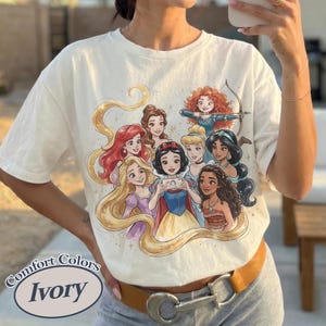 Op de afbeelding: Ivoor kleurig T-shirt met een kleurrijke afbeelding van verschillende geanimeerde prinsessen. De prinsessen worden in een groep afgebeeld, sommigen houden een telefoon vast. Het shirt heeft een ronde hals en korte mouwen. De merknaam "Comfort Colors" en de kleur "Ivory" staan op het shirt.