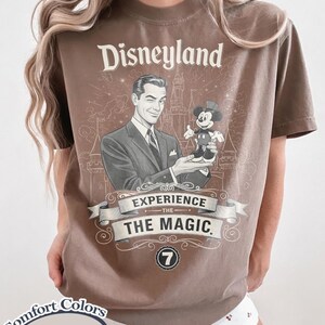 Pu&ograve; includere: T-shirt color espresso con grafica vintage di Disneyland. Il design include un uomo che tiene in mano Topolino, la scritta "Disneyland" e "Experience The Magic". La maglietta &egrave; di Comfort Colors.
