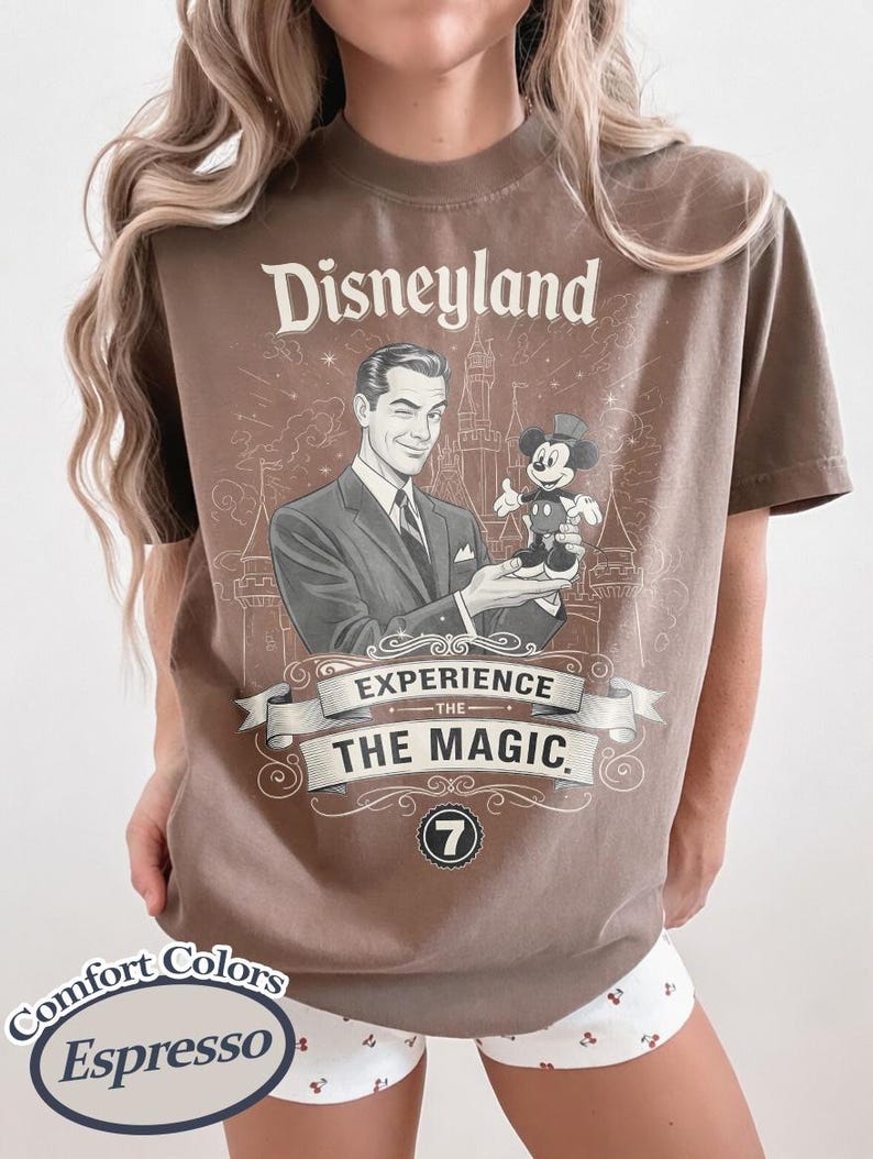 Pu&ograve; includere: T-shirt color espresso con grafica vintage di Disneyland. Il design include un uomo che tiene in mano Topolino, la scritta "Disneyland" e "Experience The Magic". La maglietta &egrave; di Comfort Colors.