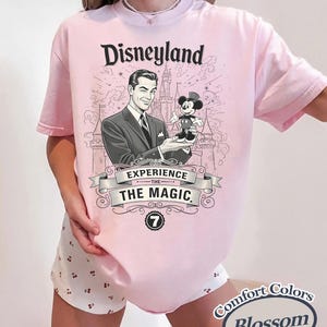Pu&ograve; includere: T-shirt rosa chiaro con grafica Disneyland. La grafica presenta un'illustrazione vintage di un uomo che tiene in mano una figura di Topolino, con la scritta "Disneyland Experience the Magic". La maglietta ha anche il logo Comfort Colors Blossom.