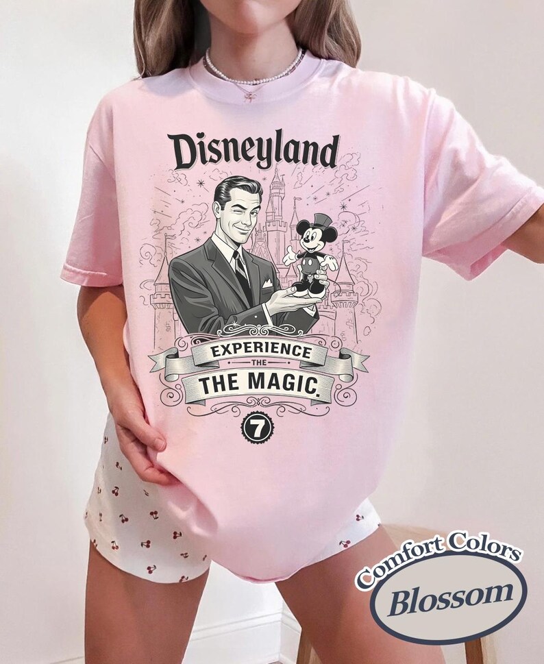 Pu&ograve; includere: T-shirt rosa chiaro con grafica Disneyland. La grafica presenta un'illustrazione vintage di un uomo che tiene in mano una figura di Topolino, con la scritta "Disneyland Experience the Magic". La maglietta ha anche il logo Comfort Colors Blossom.