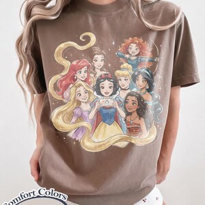 Op de afbeelding: Espresso-kleurig T-shirt met een aquarel illustratie van geanimeerde prinsessen. De prinsessen zijn omringd door fonkelende sterren en stromend haar. Op het shirt staan de woorden "Comfort Colors" en "Espresso".