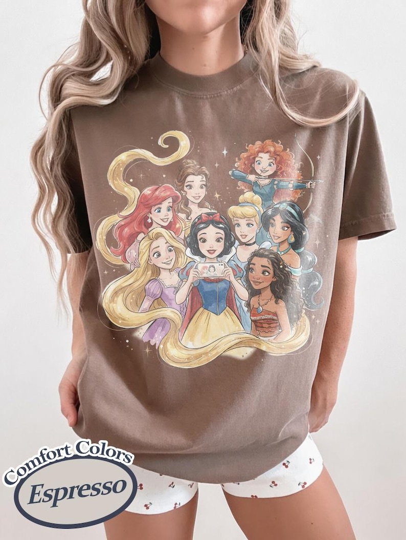 Op de afbeelding: Espresso-kleurig T-shirt met een aquarel illustratie van geanimeerde prinsessen. De prinsessen zijn omringd door fonkelende sterren en stromend haar. Op het shirt staan de woorden "Comfort Colors" en "Espresso".