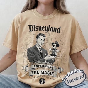 Pu&ograve; includere: T-shirt color senape con grafica vintage di Disneyland. Il design include un uomo che tiene una figura di Topolino, le parole "Disneyland" e "Experience the Magic". La maglietta ha un'estetica classica e retr&ograve;.