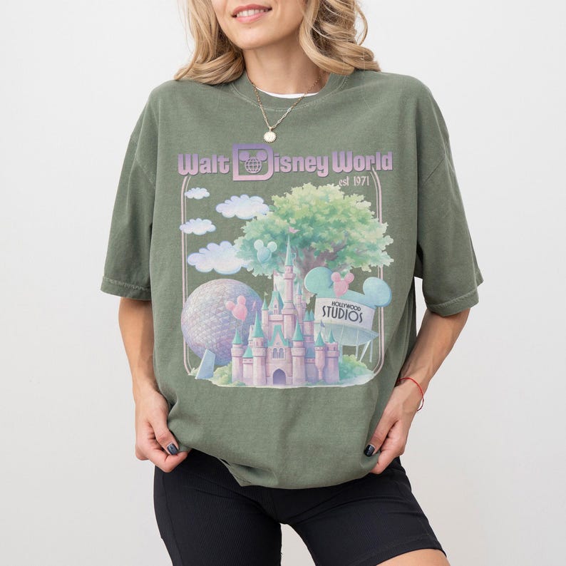 Puede incluir: Camiseta verde oliva con un dise&ntilde;o de Walt Disney World, que incluye un castillo, Spaceship Earth y nubes. El texto "Walt Disney World est 1971" est&aacute; impreso encima del dise&ntilde;o. La camiseta tiene mangas cortas y un corte holgado.