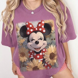 Puede incluir: Una camiseta de manga corta de color malva con un estampado gr&aacute;fico de Minnie Mouse con un vestido de lunares rojos y blancos y un lazo a juego, rodeada de girasoles amarillos. El personaje tiene una expresi&oacute;n sonriente con los ojos cerrados.