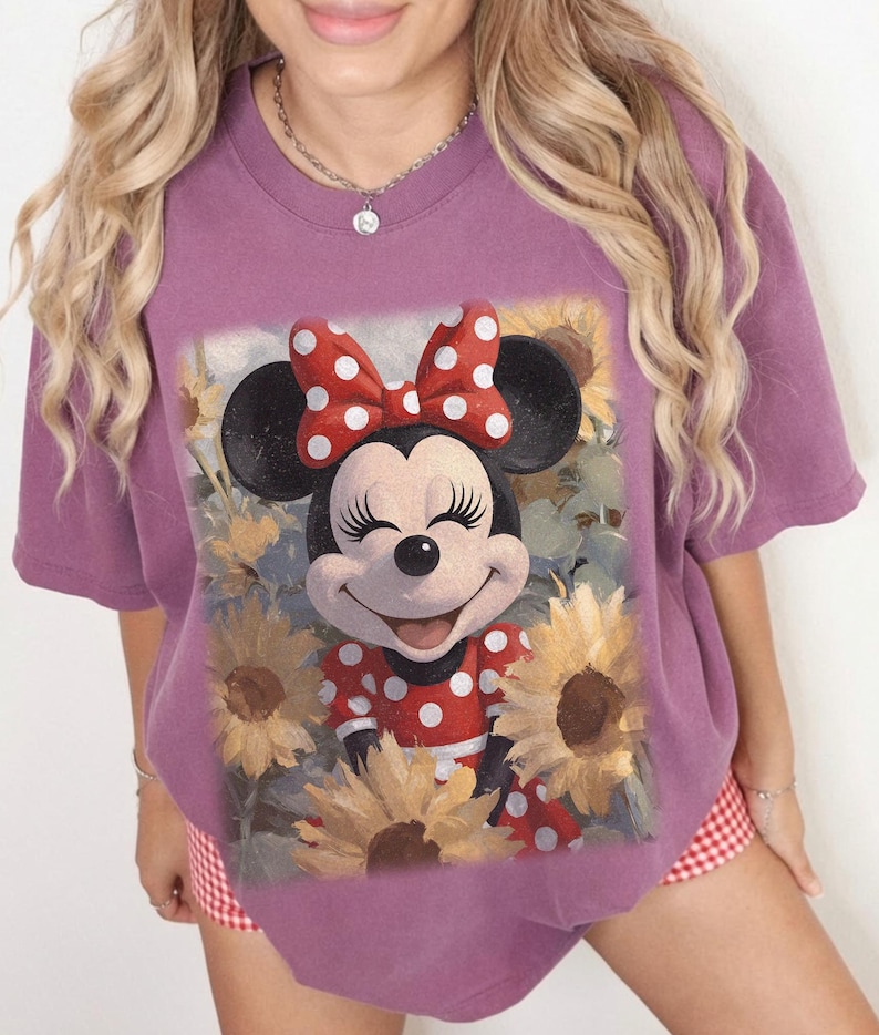 Puede incluir: Una camiseta de manga corta de color malva con un estampado gr&aacute;fico de Minnie Mouse con un vestido de lunares rojos y blancos y un lazo a juego, rodeada de girasoles amarillos. El personaje tiene una expresi&oacute;n sonriente con los ojos cerrados.