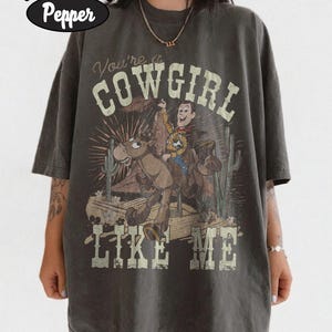 May include: A charcoal gray short-sleeve t-shirt featuring a graphic of a cowboy riding a horse in a desert landscape with cacti. The text reads "You're a COWGIRL LIKE ME" in a distressed font. The shirt is labeled as Comfort Colors Pepper.