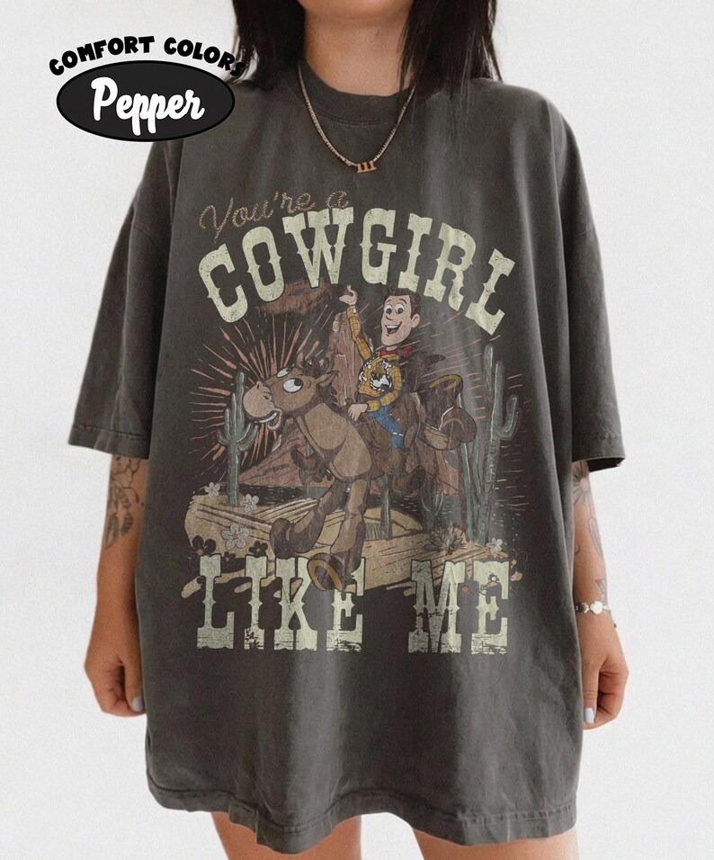 May include: A charcoal gray short-sleeve t-shirt featuring a graphic of a cowboy riding a horse in a desert landscape with cacti. The text reads "You're a COWGIRL LIKE ME" in a distressed font. The shirt is labeled as Comfort Colors Pepper.