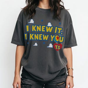 May include: A charcoal gray short-sleeve t-shirt featuring the yellow text I KNEW IT, I KNEW YOU with a red heart containing the letters TS. The design includes small white cloud shapes and blue stars scattered across the front of the garment.