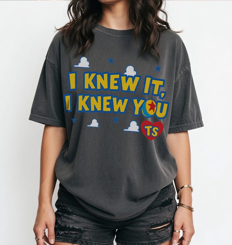 May include: A charcoal gray short-sleeve t-shirt featuring the yellow text I KNEW IT, I KNEW YOU with a red heart containing the letters TS. The design includes small white cloud shapes and blue stars scattered across the front of the garment.