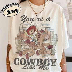 May include: An ivory t-shirt featuring a graphic print of a cartoon cowgirl riding a horse in a desert landscape with cacti. The text above reads "You're a" and below reads "COWBOY Like Me". A small oval label in the top left corner says "COMFORT COLORS IVORY".
