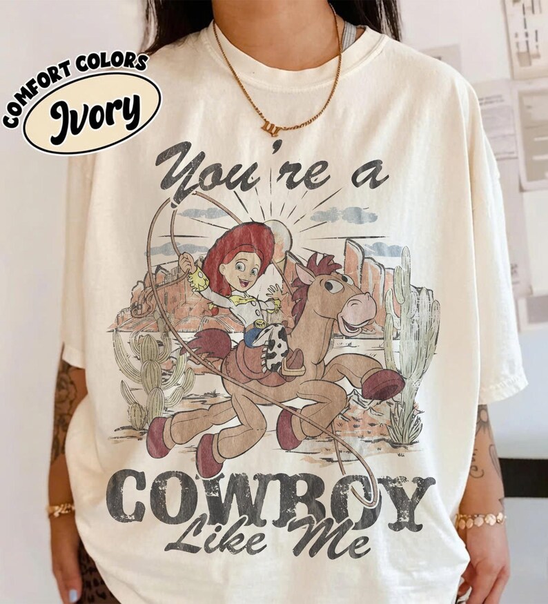 May include: An ivory t-shirt featuring a graphic print of a cartoon cowgirl riding a horse in a desert landscape with cacti. The text above reads "You're a" and below reads "COWBOY Like Me". A small oval label in the top left corner says "COMFORT COLORS IVORY".