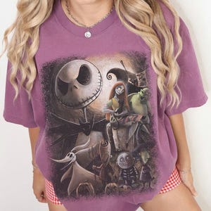 May include: A mauve short-sleeve t-shirt featuring a large graphic print of characters from The Nightmare Before Christmas, including Jack Skellington, Sally, Zero, and other figures set against a dark, textured background with pumpkins.