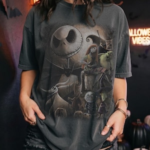 May include: A charcoal gray short-sleeve t-shirt featuring a large graphic print of characters from The Nightmare Before Christmas, including Jack Skellington, Sally, Zero, and other figures set against a full moon background with dark silhouettes.