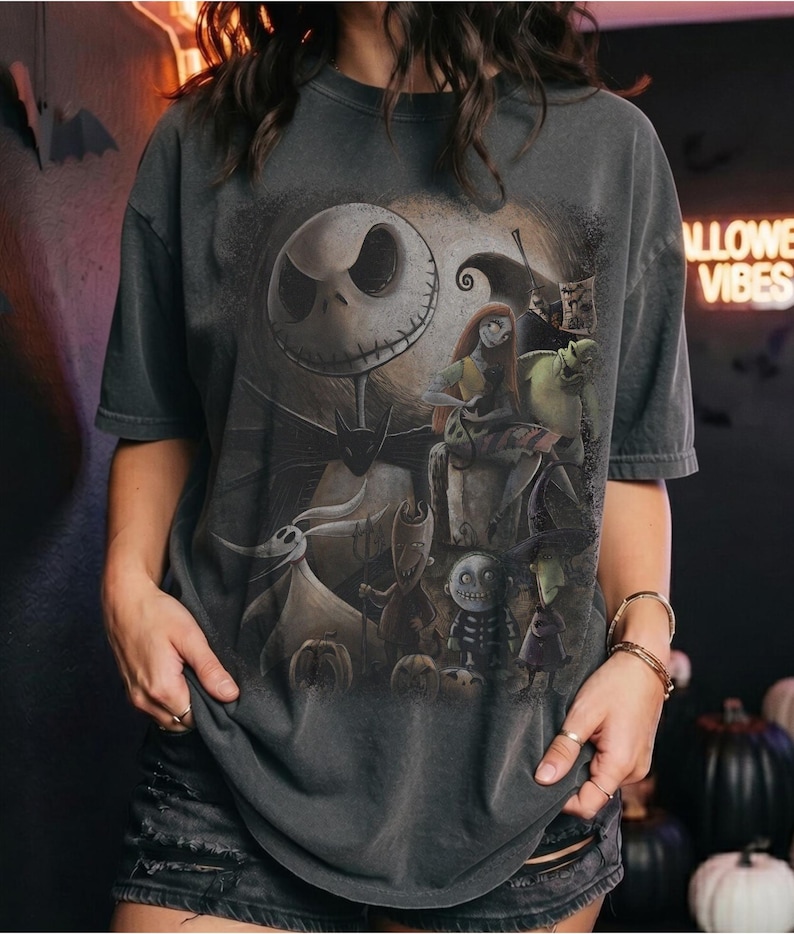 May include: A charcoal gray short-sleeve t-shirt featuring a large graphic print of characters from The Nightmare Before Christmas, including Jack Skellington, Sally, Zero, and other figures set against a full moon background with dark silhouettes.