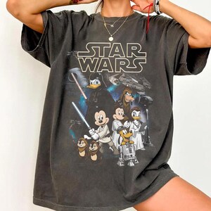 May include: Dark gray t-shirt featuring the "Star Wars" logo and a mashup of Disney characters, including Mickey Mouse, Donald Duck, and Goofy, with lightsabers and Darth Vader. The shirt has a vintage, worn look.