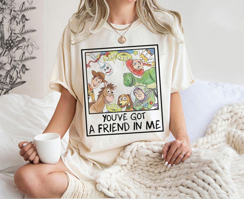 May include: Cream-colored t-shirt featuring a framed watercolor illustration of characters from the animated film "Toy Story." The image includes Woody, Buzz Lightyear, and other characters. The text "YOU'VE GOT A FRIEND IN ME" is printed below the illustration.