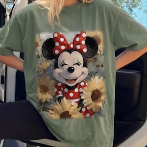 Puede incluir: Una camiseta de manga corta color verde salvia con un estampado gr&aacute;fico de Minnie Mouse con un lazo y vestido de lunares rojos y blancos, rodeada de girasoles amarillos. El personaje tiene una expresi&oacute;n sonriente con los ojos cerrados y pesta&ntilde;as largas.