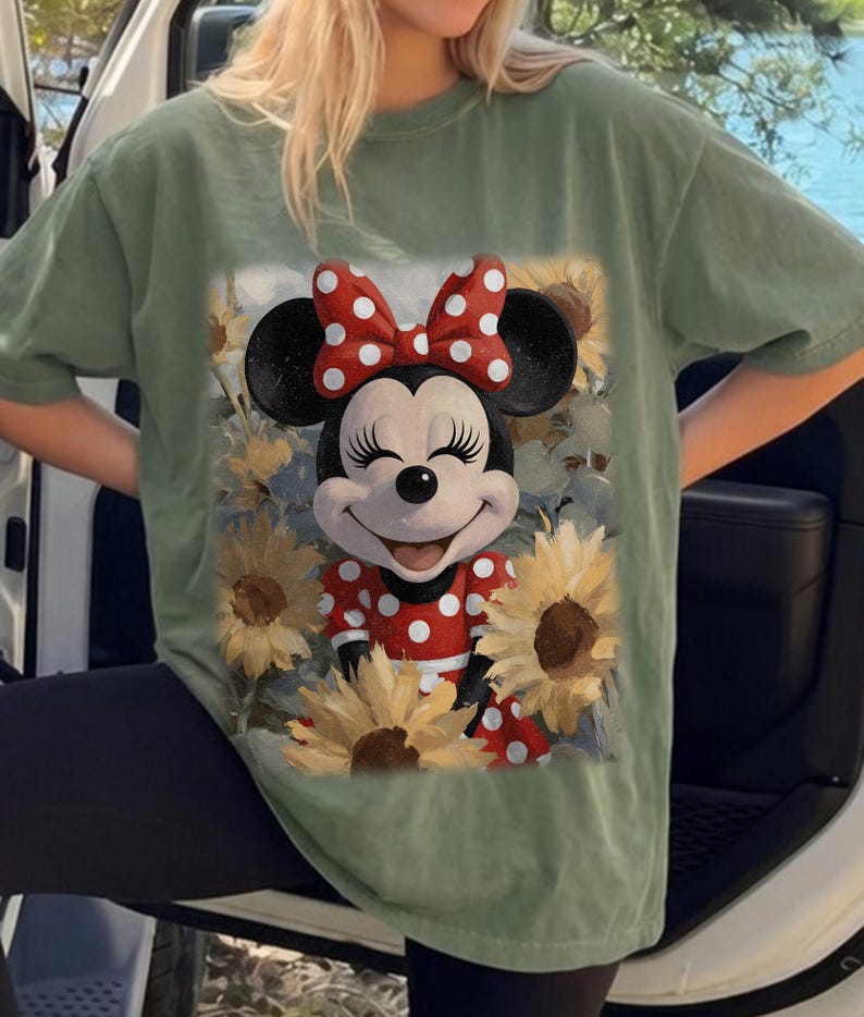 Puede incluir: Una camiseta de manga corta color verde salvia con un estampado gr&aacute;fico de Minnie Mouse con un lazo y vestido de lunares rojos y blancos, rodeada de girasoles amarillos. El personaje tiene una expresi&oacute;n sonriente con los ojos cerrados y pesta&ntilde;as largas.