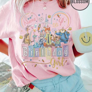 以下が含まれることがあります： 漫画のキャラクター、花火、そして「Birthday Girl」の文字が描かれたカラフルなグラフィックのピンク色のTシャツ。 デザインには数字の「4」と「Name」の文字が含まれています。 その人は水色のショートパンツを履いて、スマイリーフェイスのマグカップを持っています。