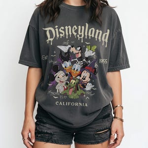 May include: A charcoal gray short-sleeve t-shirt featuring the text Disneyland Est 1955 California. The graphic displays cartoon characters including Mickey Mouse, Minnie Mouse, Donald Duck, Daisy Duck, Goofy, and Pluto in Halloween costumes.