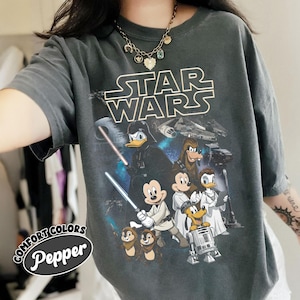 May include: Dark gray t-shirt featuring a Star Wars graphic with Disney characters. The design includes Darth Vader, Mickey Mouse as a Jedi, Donald Duck, and other characters. The shirt has the text "STAR WARS" in white and the text "COMFORT COLORS Pepper" in a circular logo.