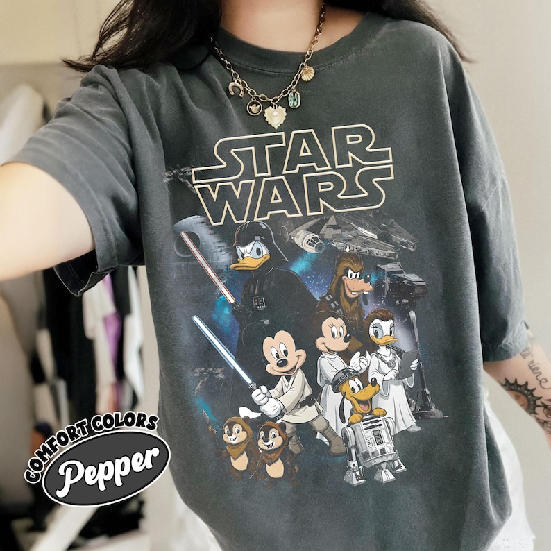May include: Dark gray t-shirt featuring a Star Wars graphic with Disney characters. The design includes Darth Vader, Mickey Mouse as a Jedi, Donald Duck, and other characters. The shirt has the text "STAR WARS" in white and the text "COMFORT COLORS Pepper" in a circular logo.