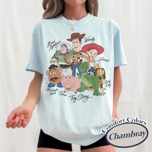Puede incluir: Camiseta azul claro con un gr&aacute;fico de personajes de Toy Story, incluyendo a Woody y Buzz Lightyear. La camiseta tiene las palabras "Toy Story" y "Comfort Colors Chambray" impresas.