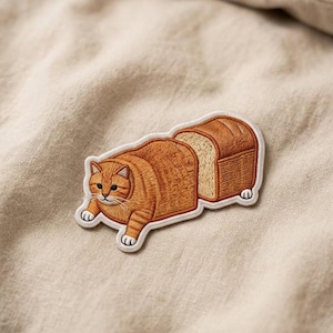 May include: Embroidered patch featuring a cartoon cat shaped like a loaf of bread. The orange tabby cat is depicted with white paws and a white outline. The bread loaf has a slice cut out, revealing the interior of the bread.