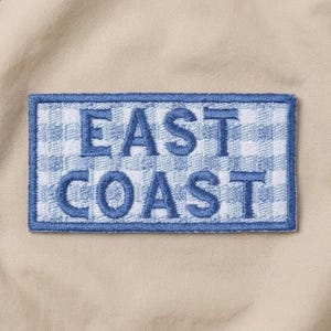 May include: Embroidered patch with the words "EAST COAST" in blue, block letters. The patch has a blue border and a light blue and white gingham pattern background. The patch is rectangular and set against a tan fabric.