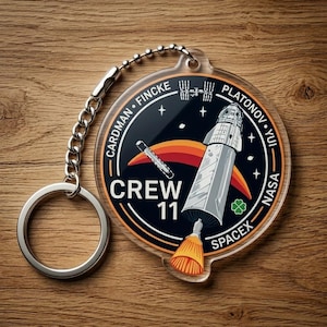May include: Clear acrylic keychain featuring a circular design with a space theme. The design includes a rocket, the text "CREW 11", and the names CARDMAN, FINCKE, PLATONOV, and YUI. The keychain has a silver chain and keyring.